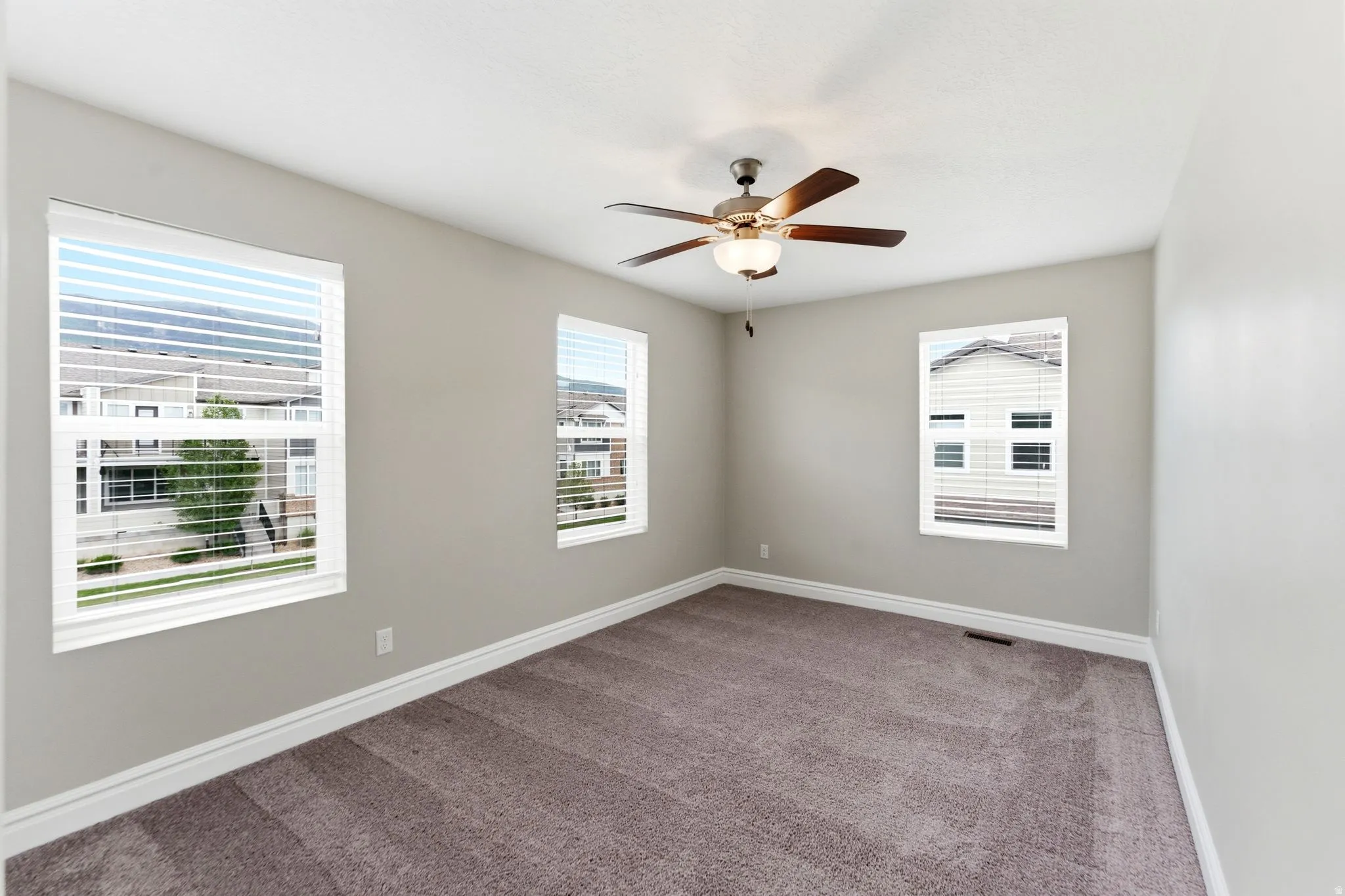 Spacious room featuring multiple windows with blinds, a ceiling fan with light fixture, neutral wall paint, and plush carpet flooring
