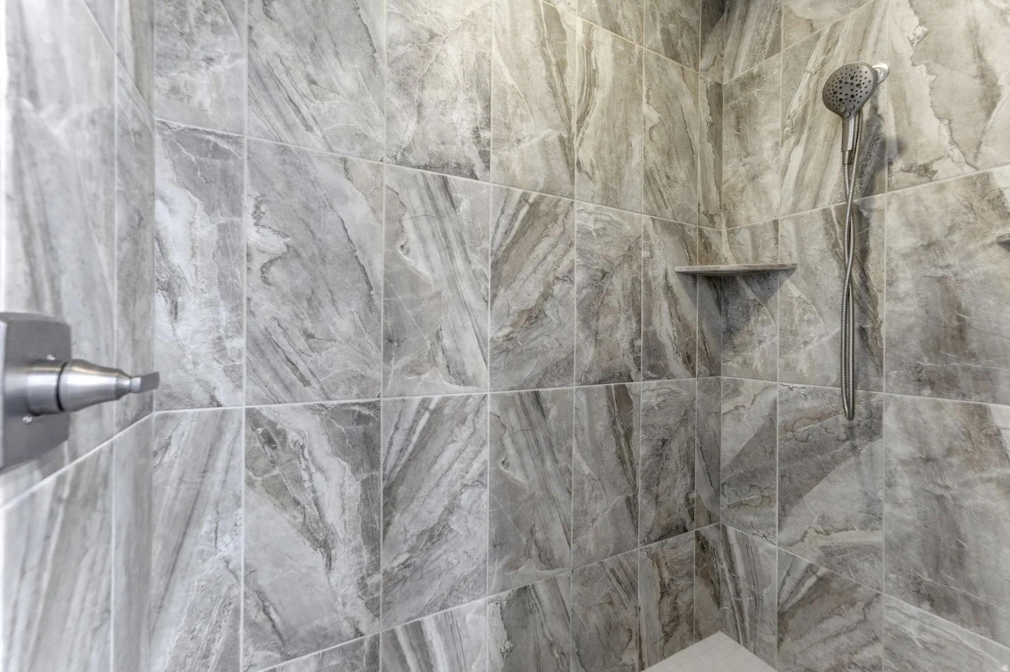 Shower enclosure featuring large format stone-look tile, a built-in corner shelf, a handheld shower fixture, a fixed shower head, and a brushed nickel control handle