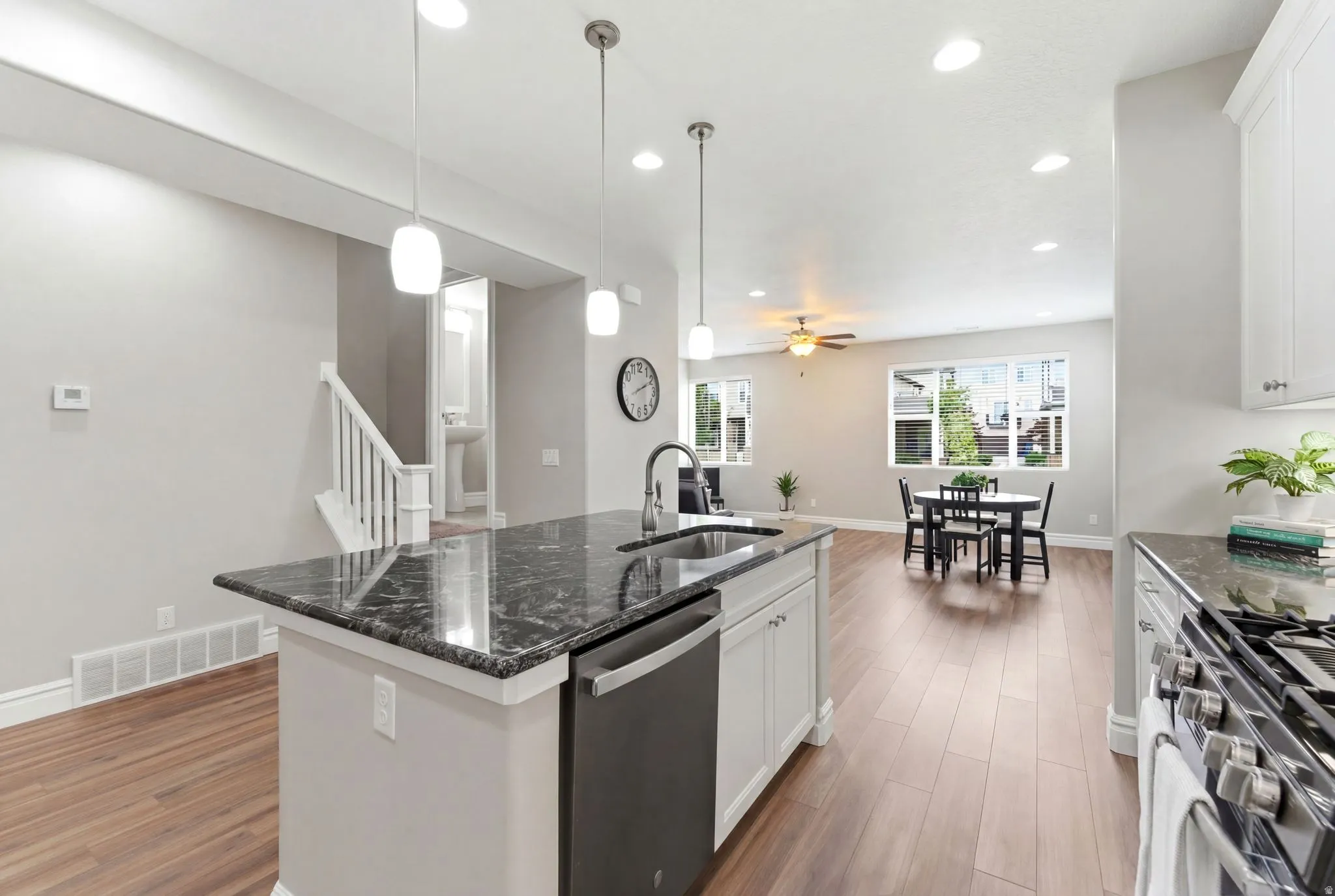 Kitchen island with dark countertop and stainless steel sink, wood-finish flooring, white cabinetry, recessed lighting, and a white staircase with wooden handrail