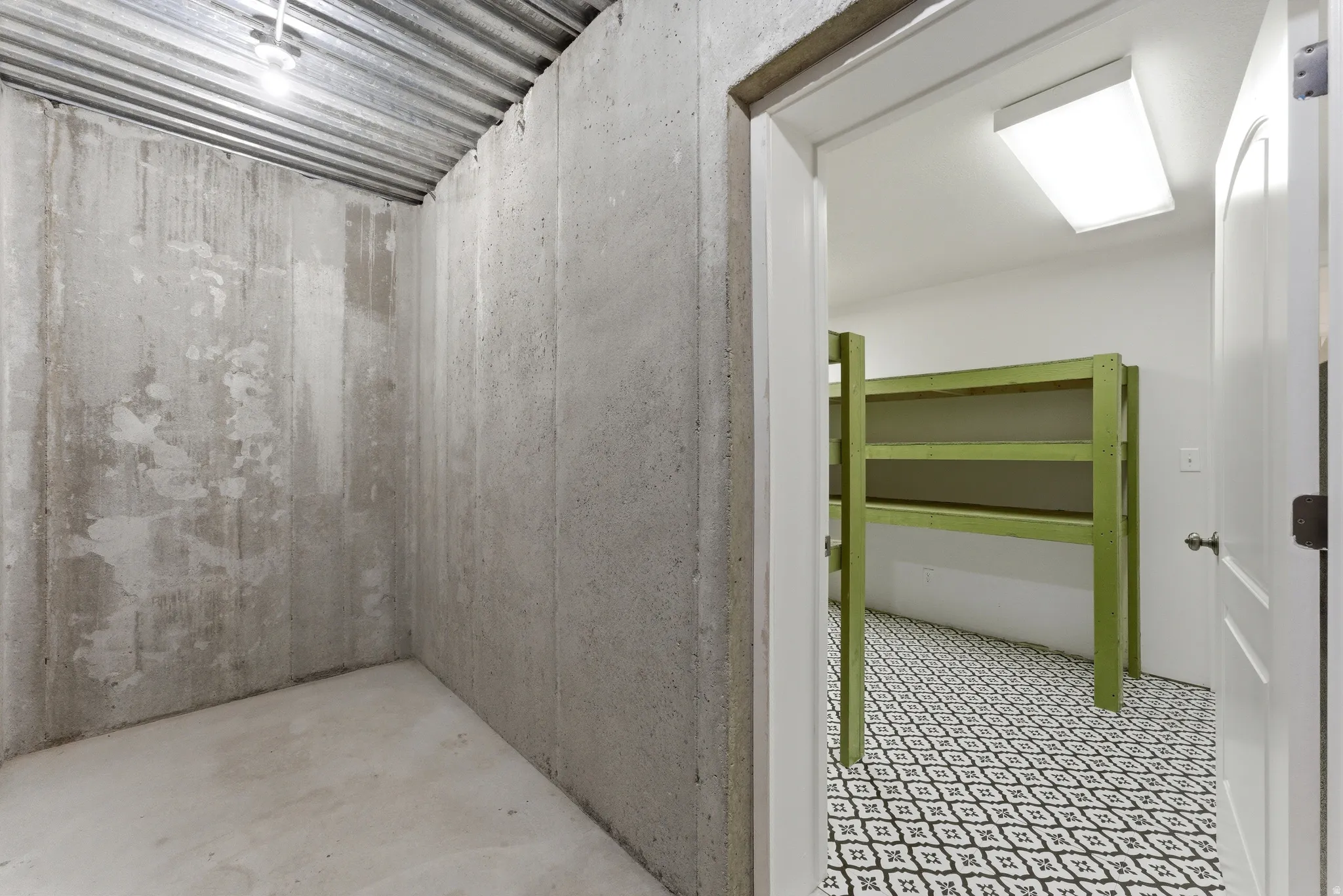 Exposed concrete walls and ceiling create a robust aesthetic, contrasting with a separate room featuring patterned floor tiling and overhead lighting