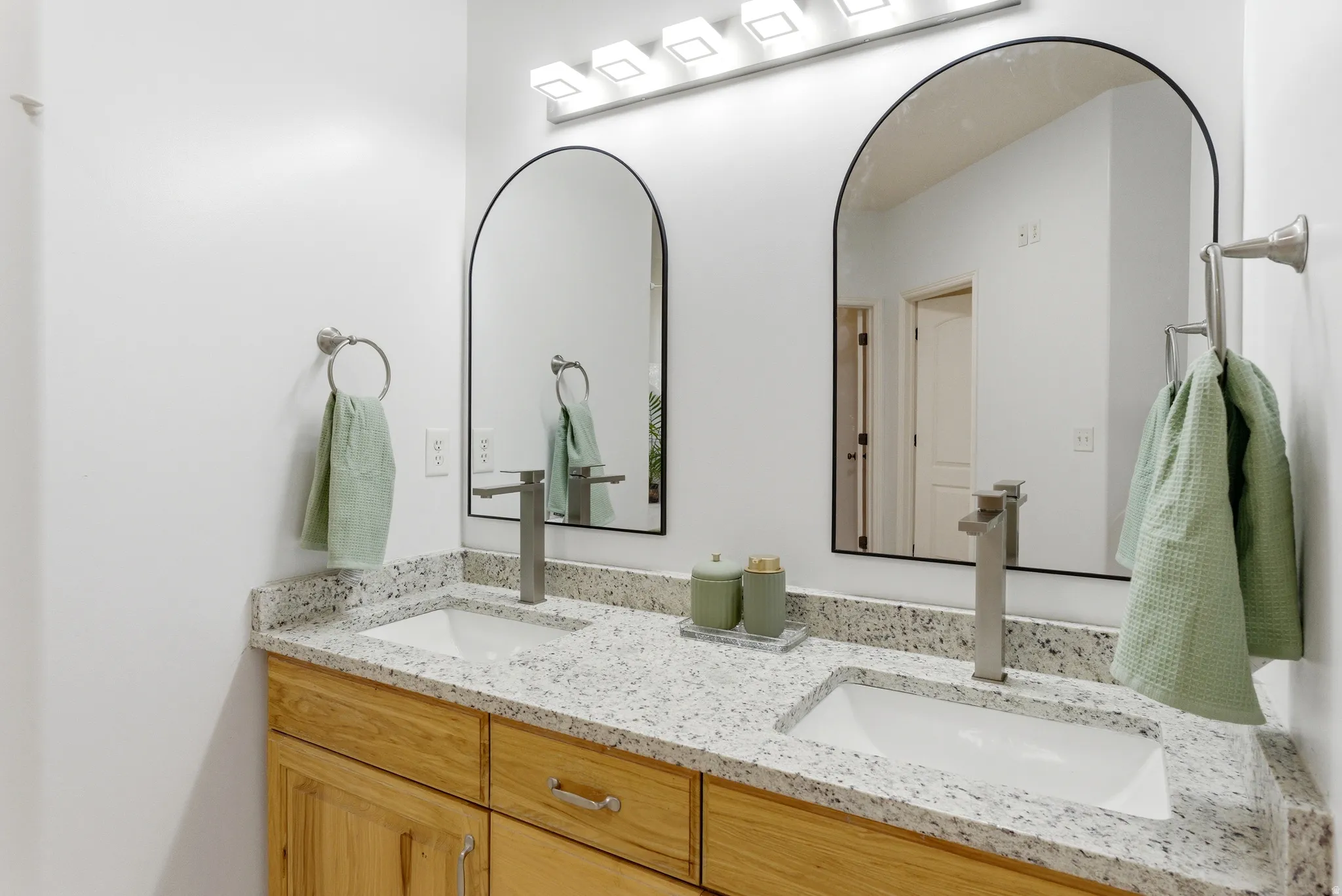 Dual vanity featuring wood-finish cabinetry, light speckled countertops, rectangular undermount sinks with contemporary faucets, and arched mirrors with black frames
