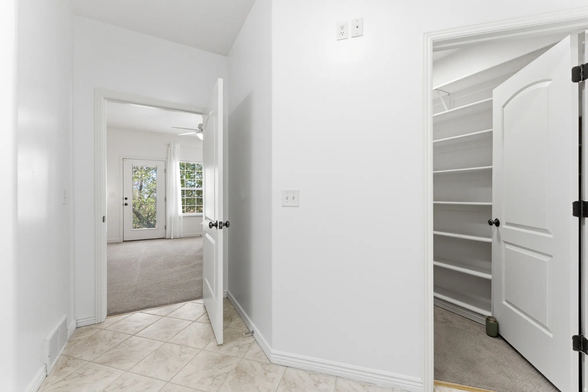 Tiled entryway featuring a built-in pantry with shelving and a doorway opening to a carpeted room with an exterior glass-paned door and window