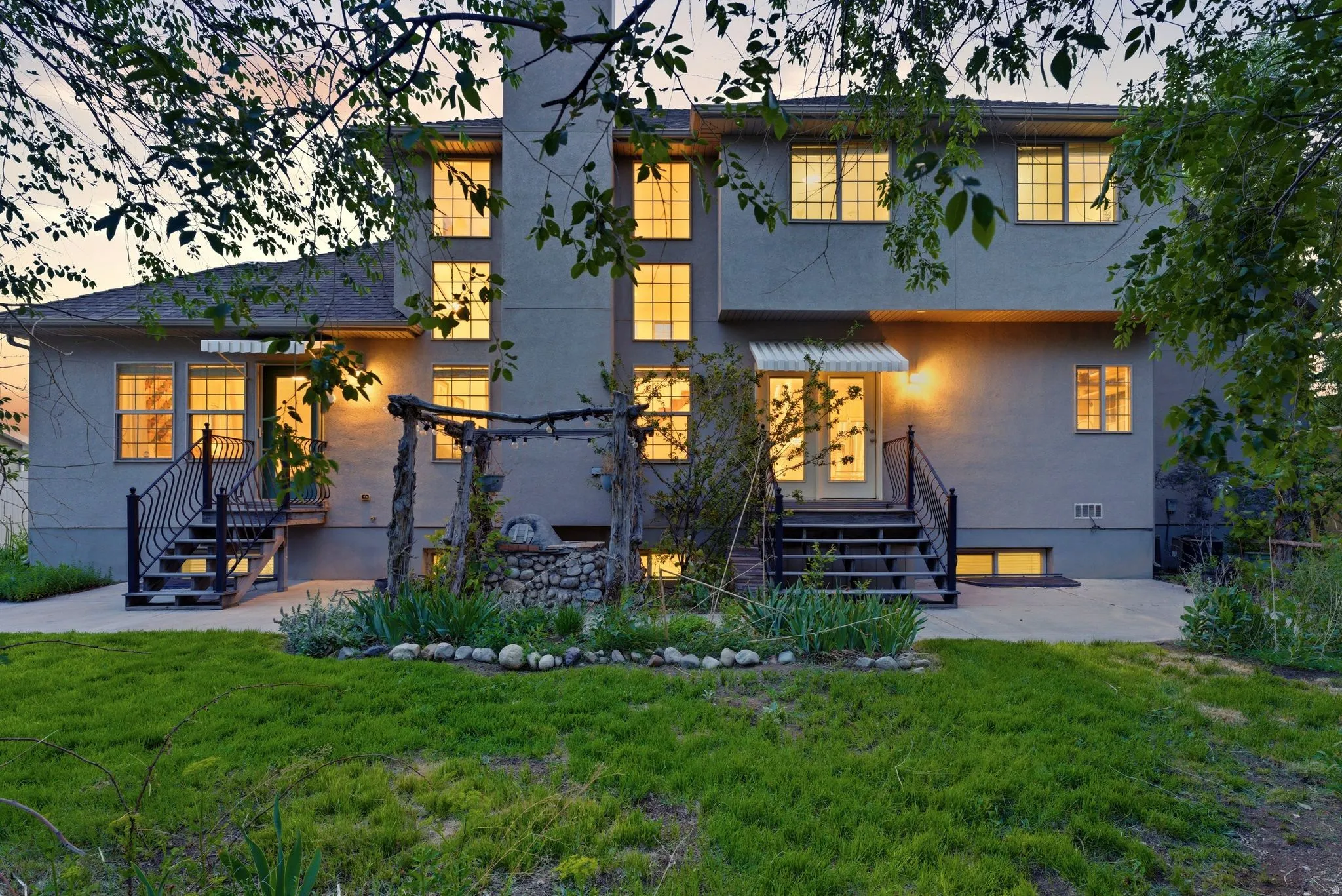 Rear exterior featuring a stucco finish, multiple windows providing natural light, two exterior staircases with wrought iron railings, a central stone-ringed garden bed, and a concrete patio area