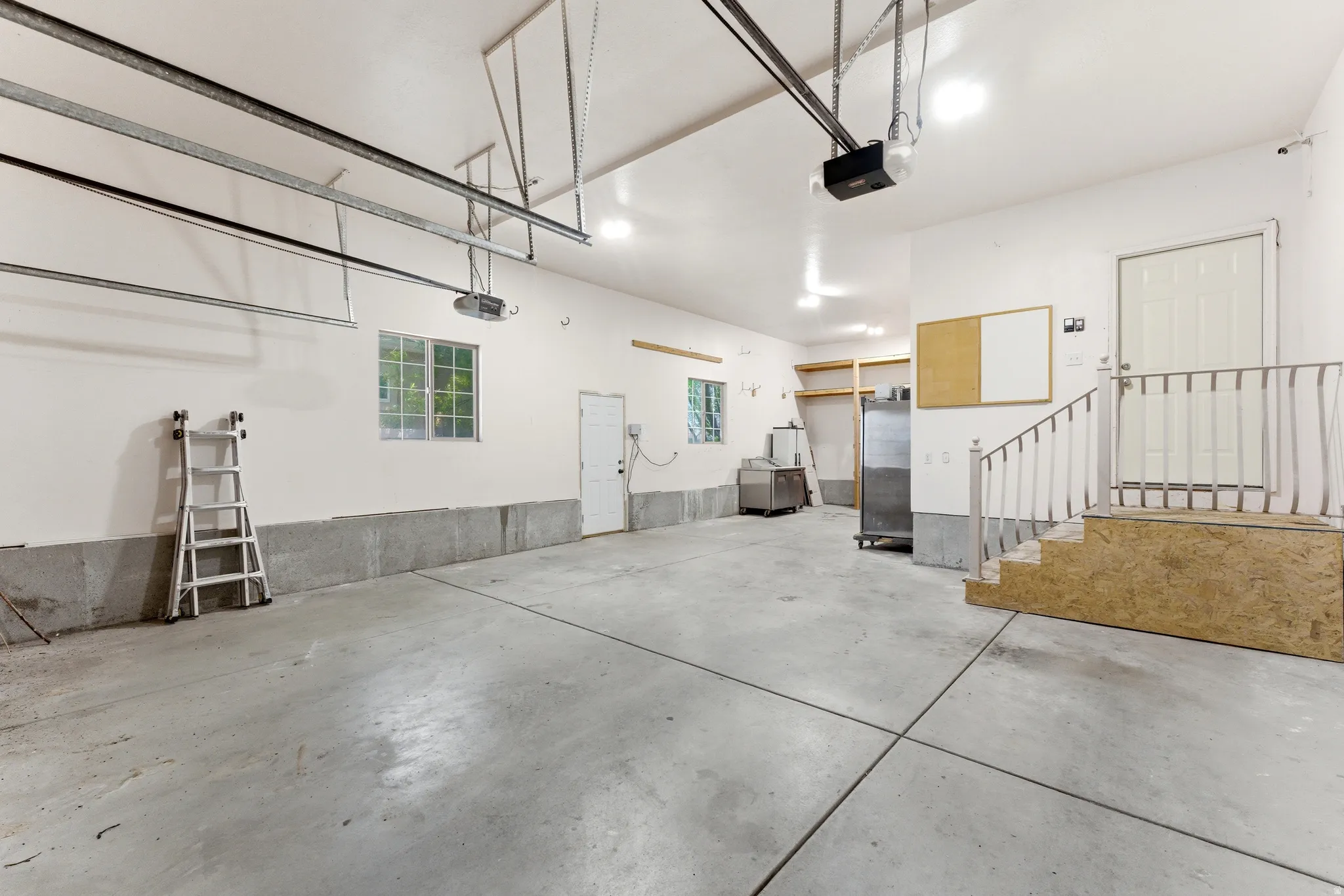 Spacious garage featuring a concrete floor, multiple windows, built-in shelving, two overhead garage door mechanisms, and an interior access door with staircase