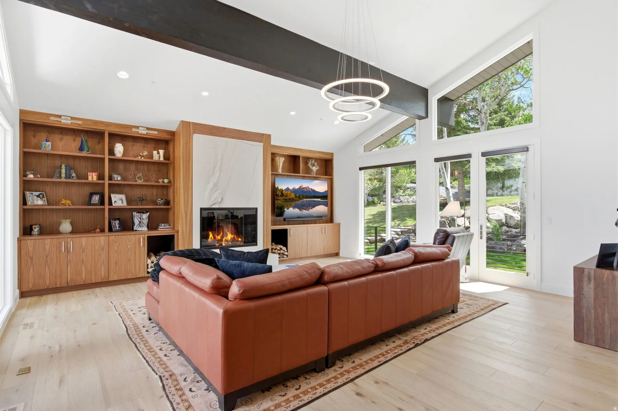 Spacious living area featuring a stone fireplace surround, built-in shelving, wood-finish flooring, vaulted ceilings with exposed beams, and multiple large windows