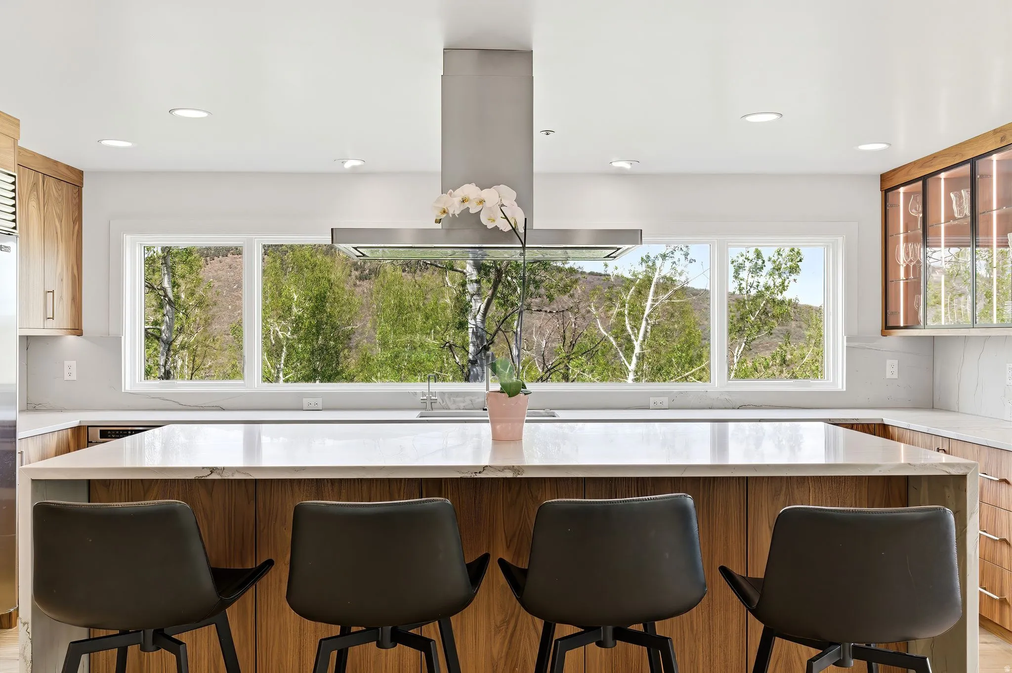 Kitchen featuring a waterfall island with stone countertop, wood-finish cabinetry, stainless steel range hood, recessed lighting, and a panoramic window overlooking natural scenery