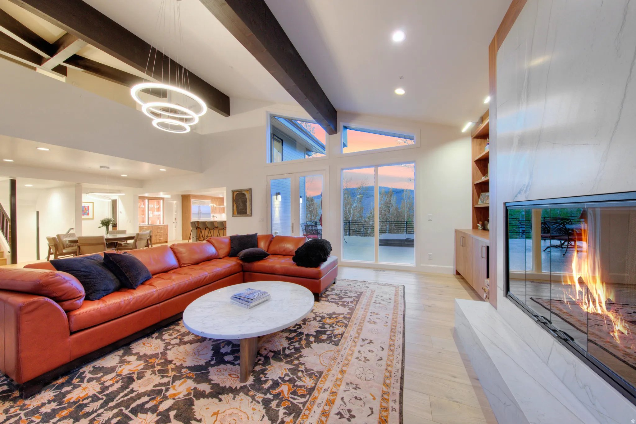Spacious living area featuring a vaulted ceiling with exposed wood beams, a contemporary linear fireplace, built-in shelving, and light wood-finish flooring