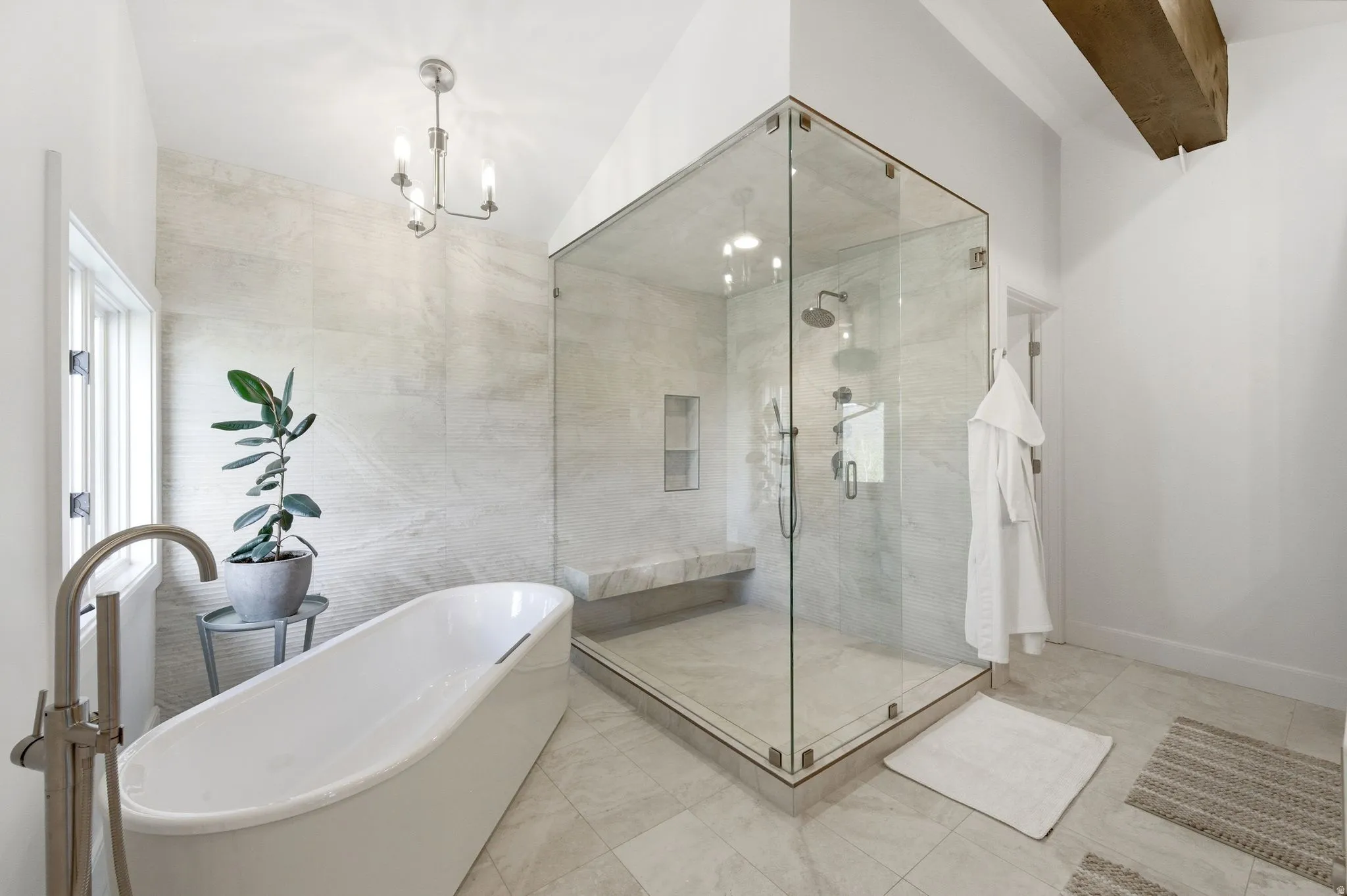 Spacious bathroom featuring a freestanding soaking tub, a large glass-enclosed shower with a built-in bench, light-toned tile flooring, and a exposed wood beam accent