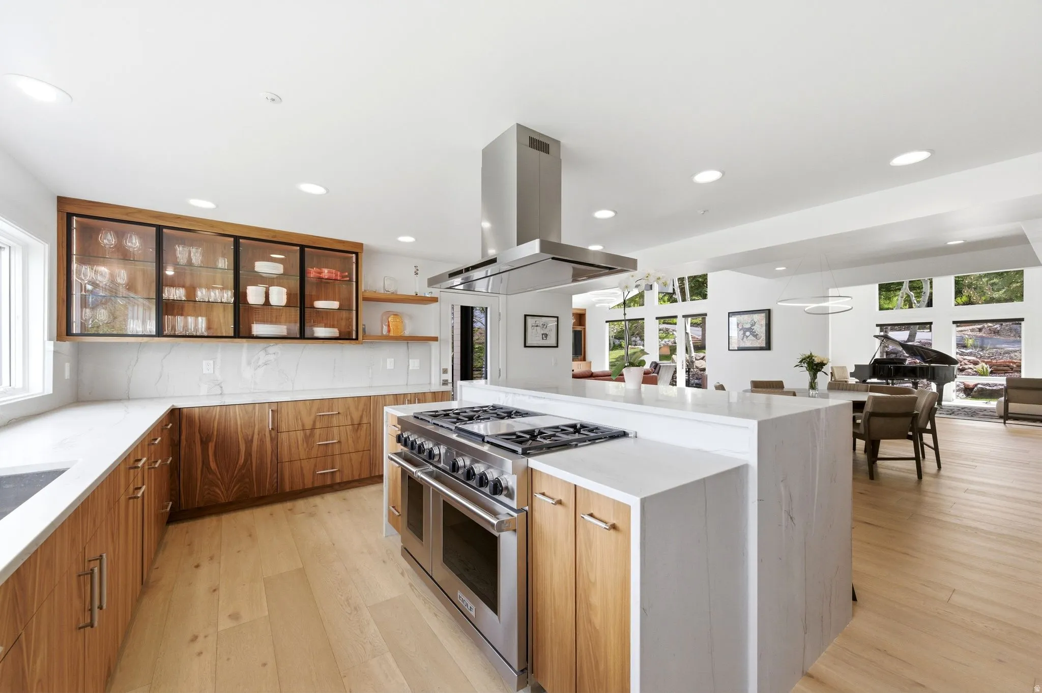 Gourmet kitchen featuring a large central island with a gas range and stainless steel hood, light wood-finish cabinetry, white countertops, and a white tile backsplash