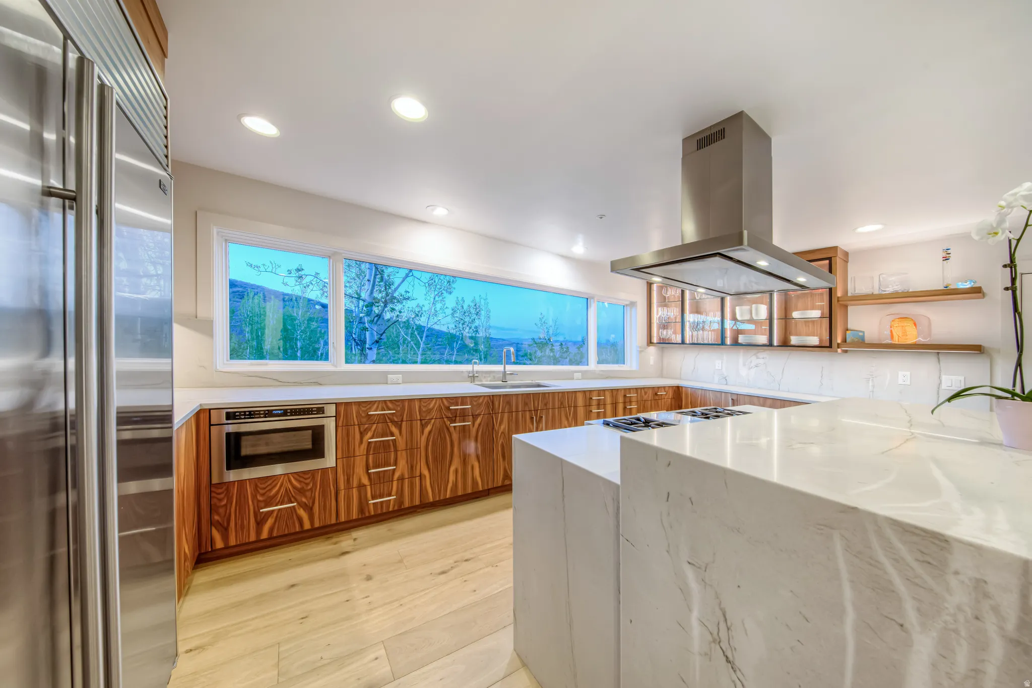 Modern kitchen featuring a large waterfall-edge island with an integrated cooktop, stainless steel appliances, extensive wood-finish cabinetry, a wide horizontal window, and recessed ceiling lighting