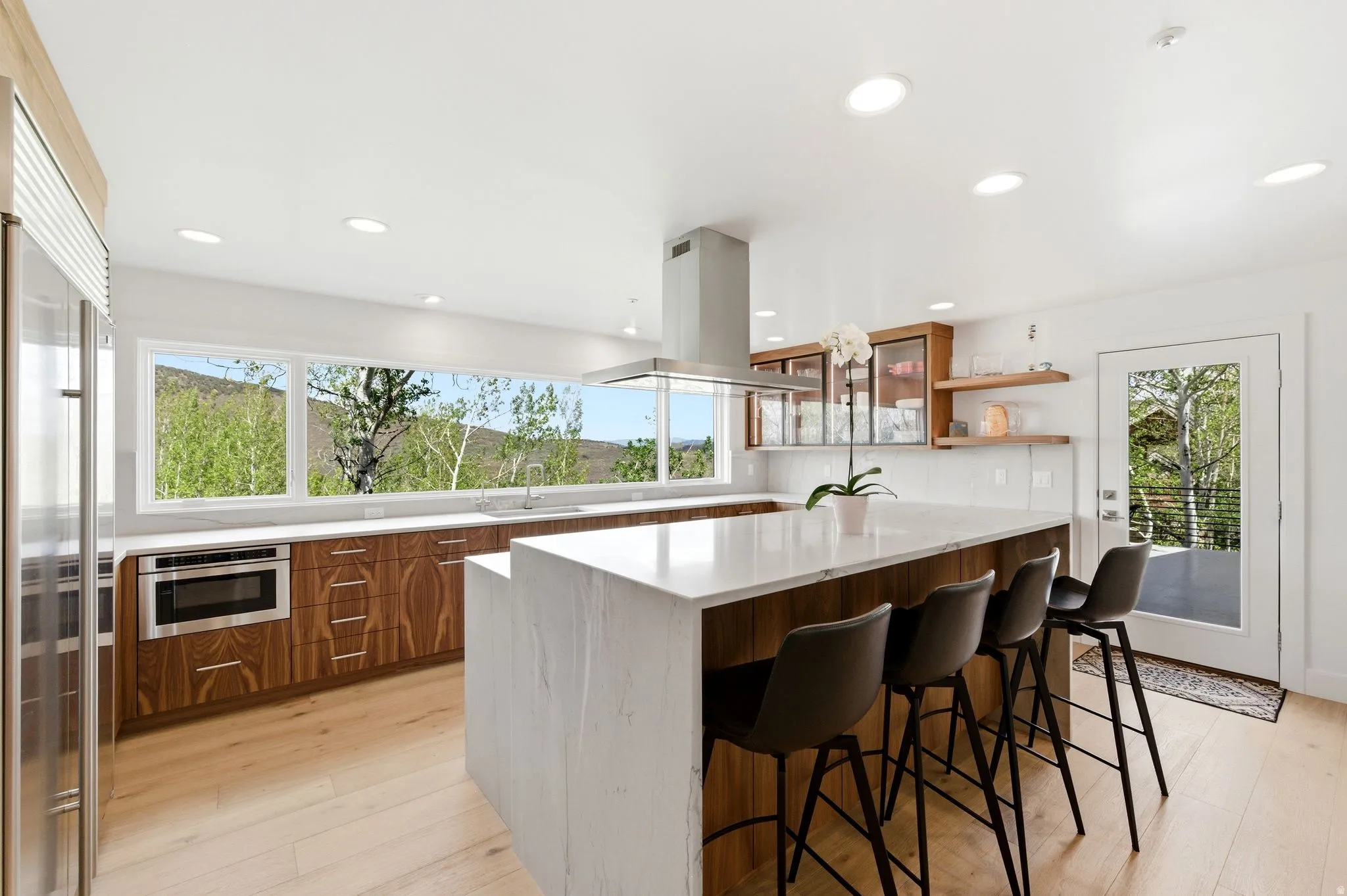Modern kitchen featuring a large waterfall island with seating, a stainless steel range hood, built-in microwave, and wood-finish cabinetry
