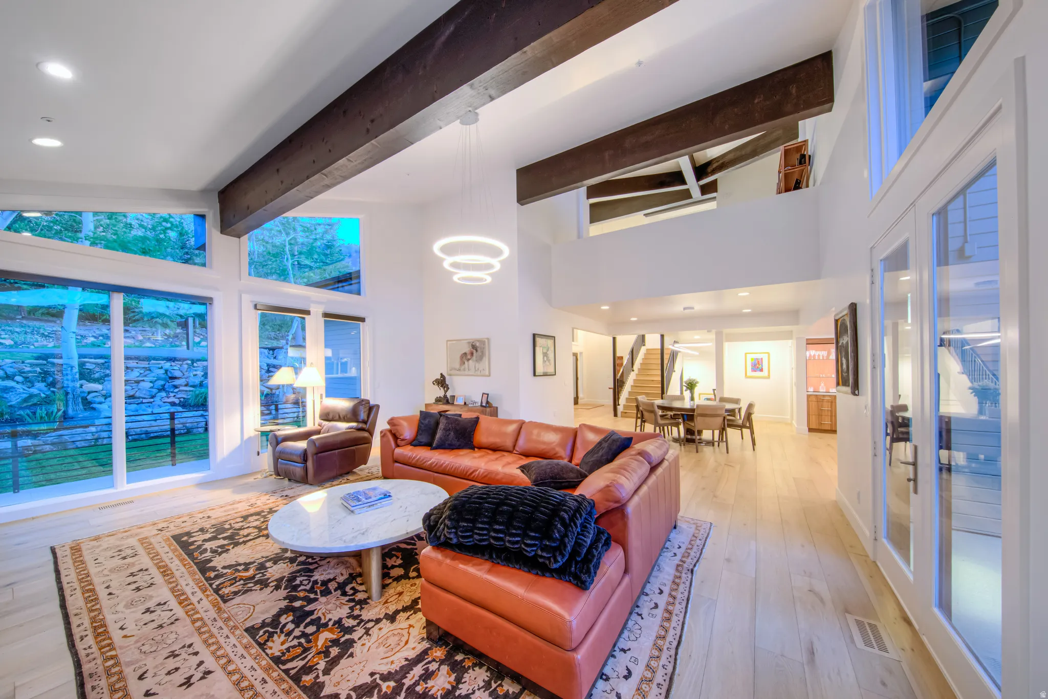 Vaulted ceilings with exposed wood beams define this expansive room, featuring large windows, wood-finish flooring, and a contemporary chandelier