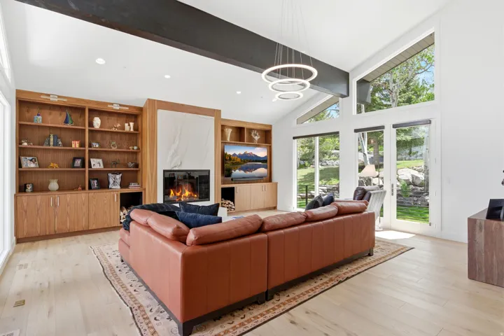 Spacious living area featuring a stone fireplace surround, built-in shelving, wood-finish flooring, vaulted ceilings with exposed beams, and multiple large windows