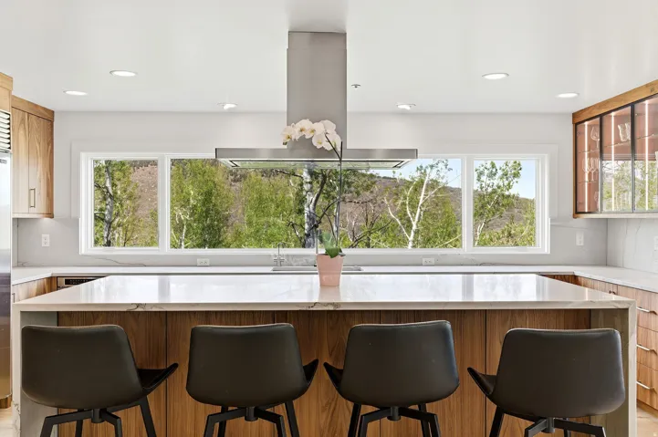 Kitchen featuring a waterfall island with stone countertop, wood-finish cabinetry, stainless steel range hood, recessed lighting, and a panoramic window overlooking natural scenery