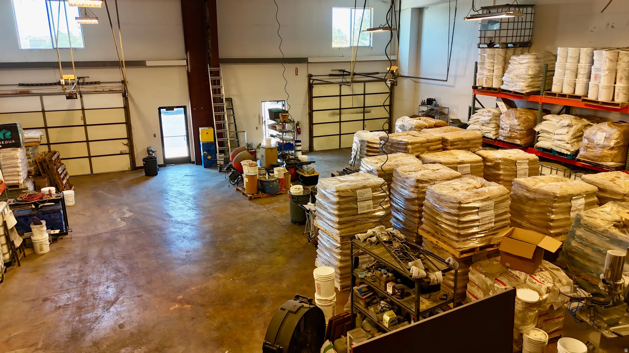 Spacious commercial interior featuring high ceilings, exposed metal beams, multiple overhead garage doors, an industrial concrete floor, and ample storage shelving