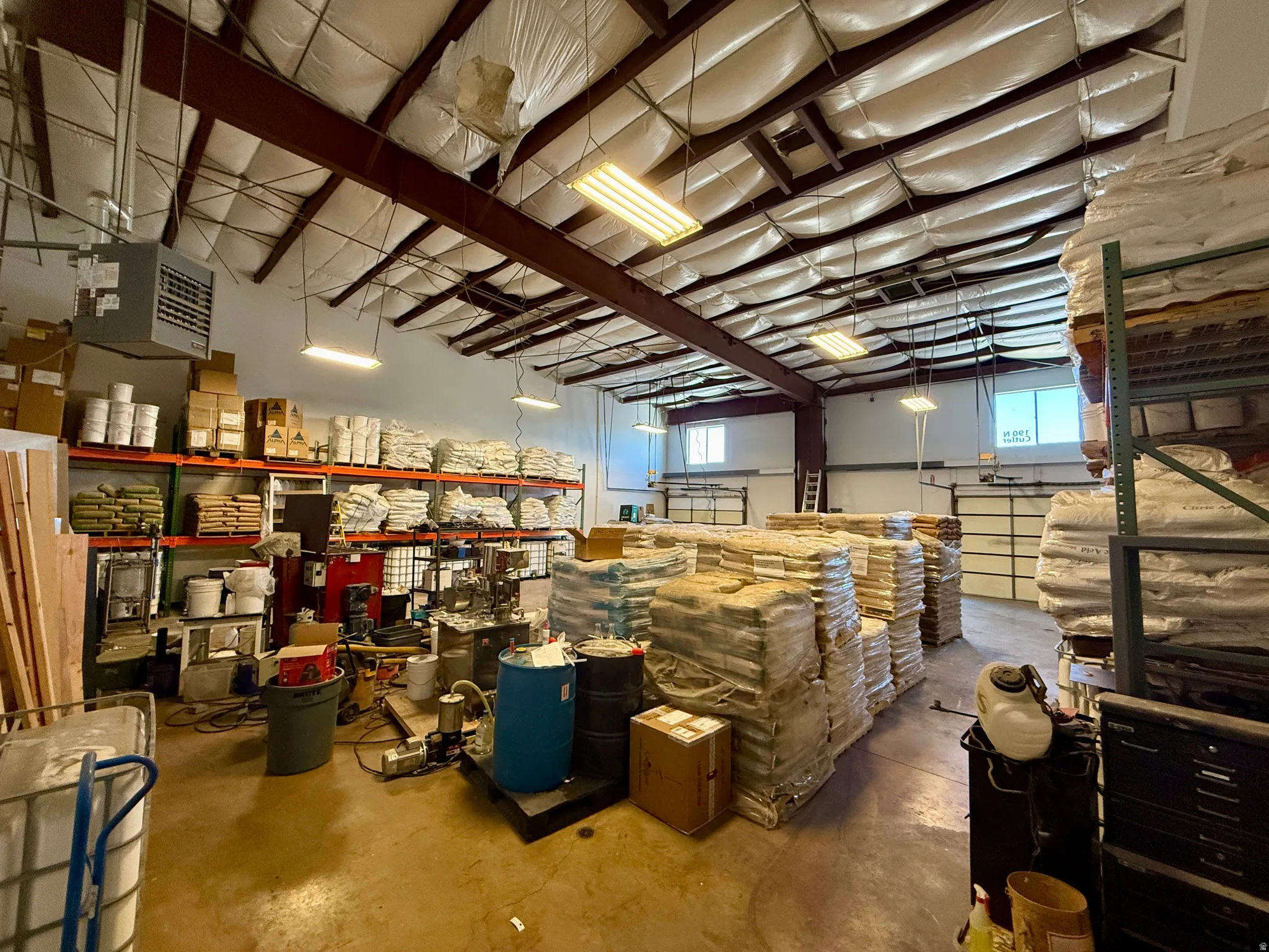 Spacious industrial interior featuring an exposed beam ceiling with insulation, multiple fluorescent light fixtures, polished concrete flooring, and an overhead garage door