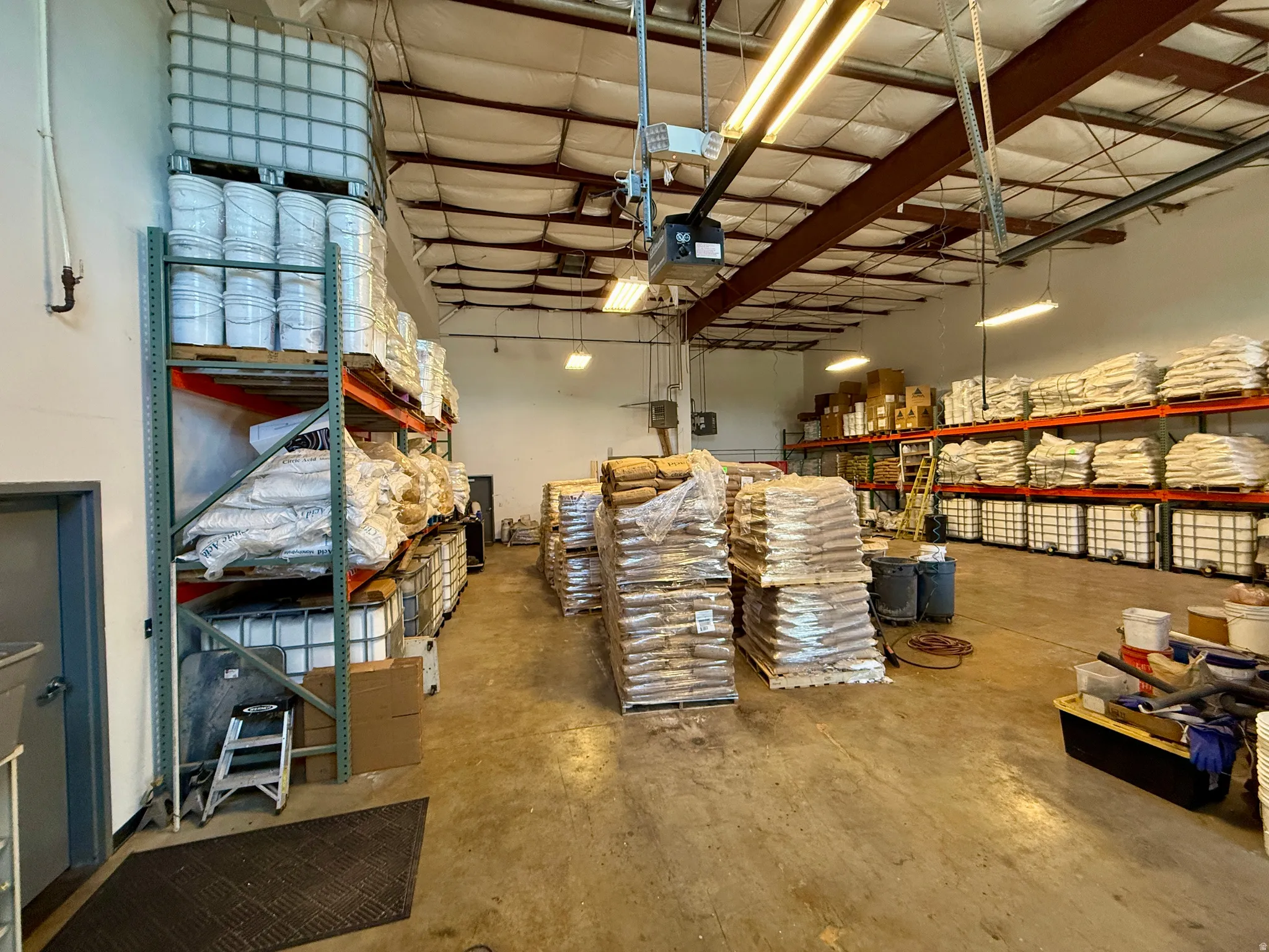 Expansive warehouse space featuring high ceilings with exposed metal beams, concrete flooring, and extensive wall-mounted shelving systems