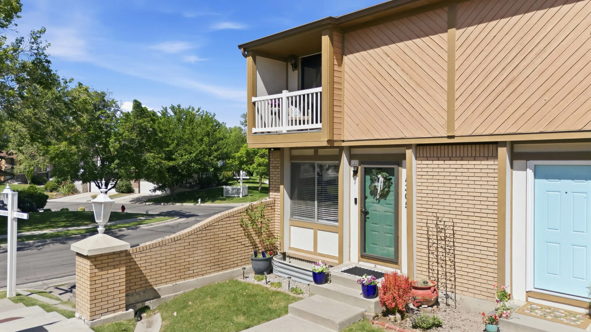Exterior featuring a second-story balcony, horizontal and diagonal siding, a green front door with sidelight, mature landscaping, and a brick half-wall