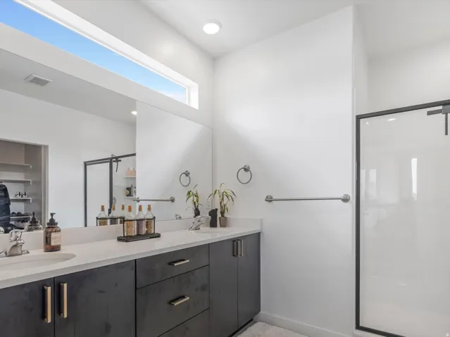Dual vanity with dark cabinetry and light countertops, a walk-in shower with a black-framed glass enclosure, a wide mirror, and a transom window