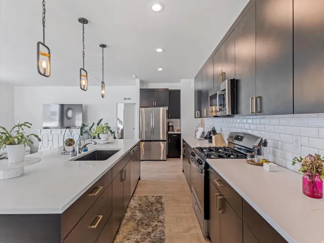 Modern kitchen featuring an expansive center island with an undermount sink, stainless steel appliances, dark wood-finish cabinetry with gold-tone hardware, white countertops, and a white subway tile backsplash