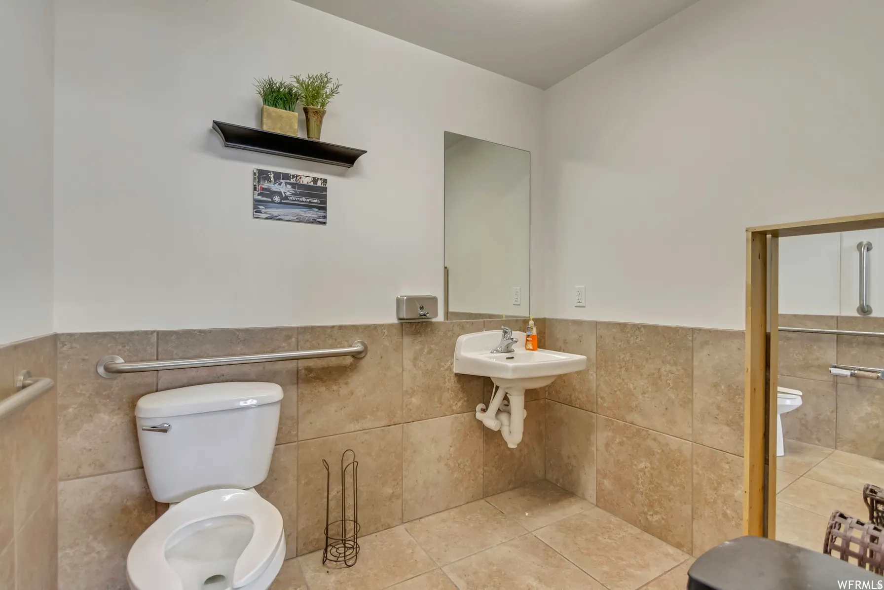 Restroom with floor-to-ceiling tile, wall-mounted sink, and a commode with grab bars