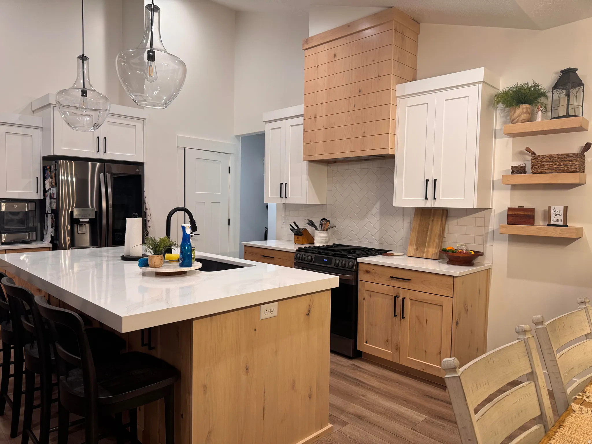 Modern kitchen featuring a large central island with a built-in sink and seating, two-tone cabinetry with light wood-finish bases and white upper cabinets, a custom wood-finish range hood, stainless steel appliances, and wood-finish flooring