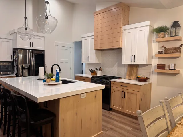 Modern kitchen featuring a large central island with a built-in sink and seating, two-tone cabinetry with light wood-finish bases and white upper cabinets, a custom wood-finish range hood, stainless steel appliances, and wood-finish flooring
