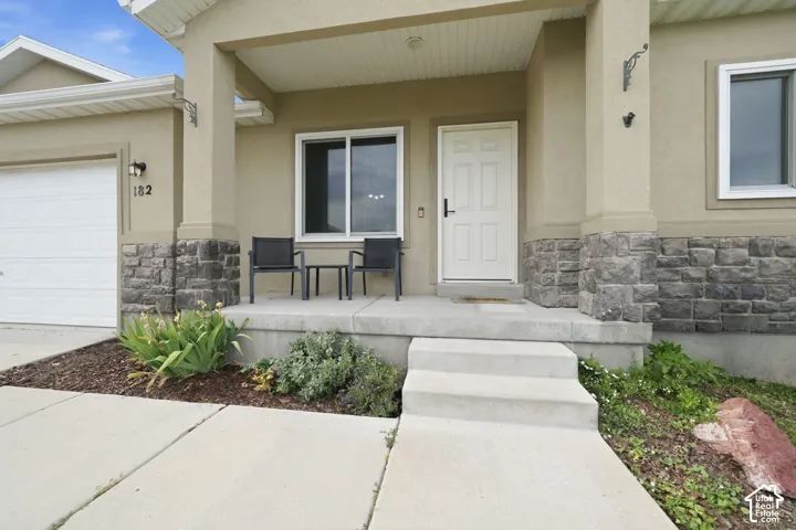 Front exterior featuring a covered porch, stone veneer accents, stucco siding, a panel garage door, and mature landscaping