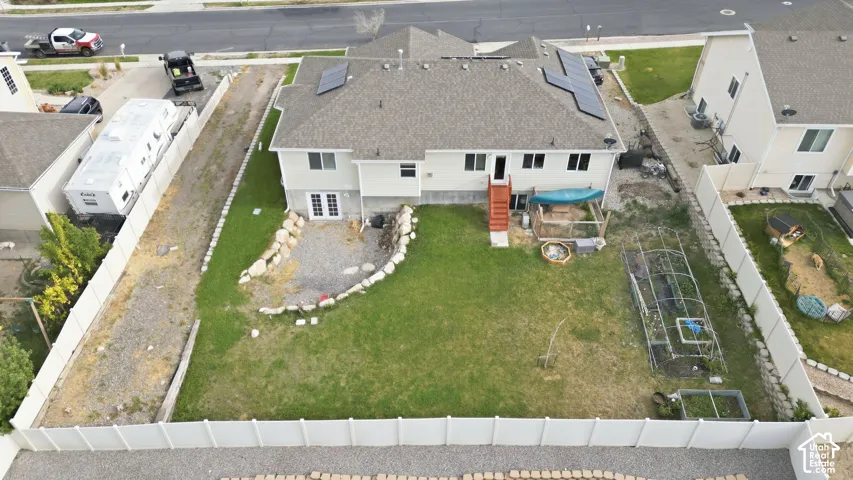 Rear exterior featuring multiple solar panels on the roof, a walk-out basement with double doors, an uncovered patio area with stone retaining walls, a grassy lawn, and a fenced perimeter