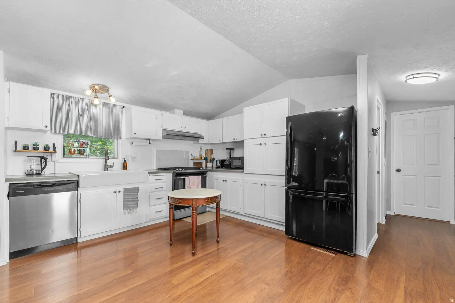 Kitchen featuring wood-finish flooring, white cabinetry, a farmhouse sink, stainless steel dishwasher, and a black refrigerator