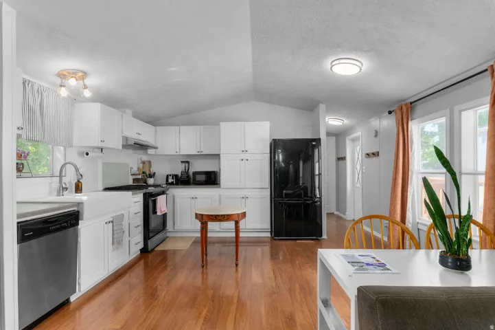 Open-concept kitchen and dining area featuring wood-finish flooring, white cabinetry, stainless steel dishwasher, and a farmhouse-style sink