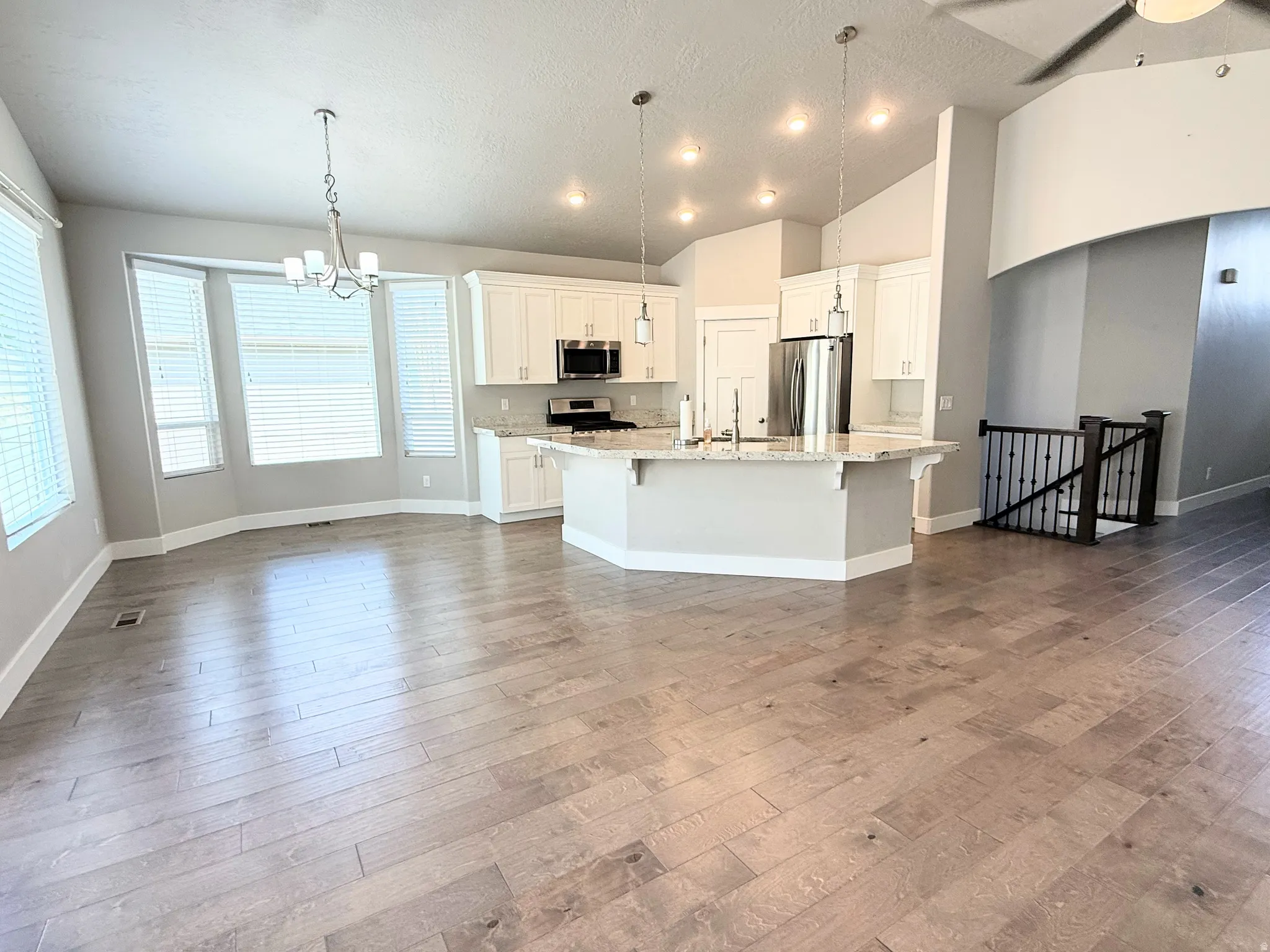 Open-concept living area featuring wood-finish flooring, a contemporary kitchen island with pendant lighting, white cabinetry, stainless steel appliances, and a bay window