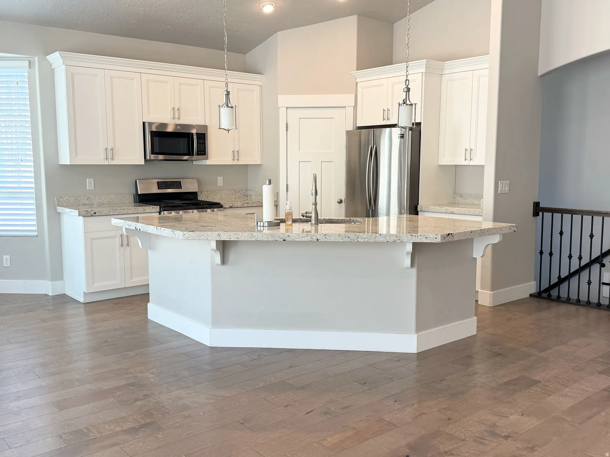 Spacious kitchen featuring an angular island with an undermount sink, pendant lighting, white cabinetry, and stainless steel appliances