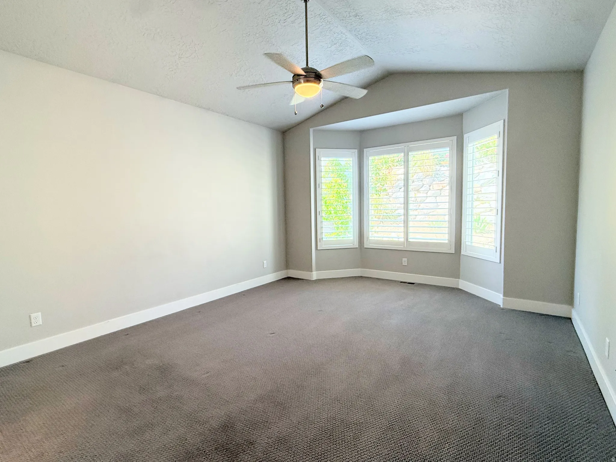 Spacious Primary Suite featuring a vaulted ceiling and a ceiling fan with integrated lighting, plantation shutters, and soft gray carpeting