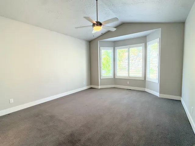 Spacious Primary Suite featuring a vaulted ceiling and a ceiling fan with integrated lighting, plantation shutters, and soft gray carpeting