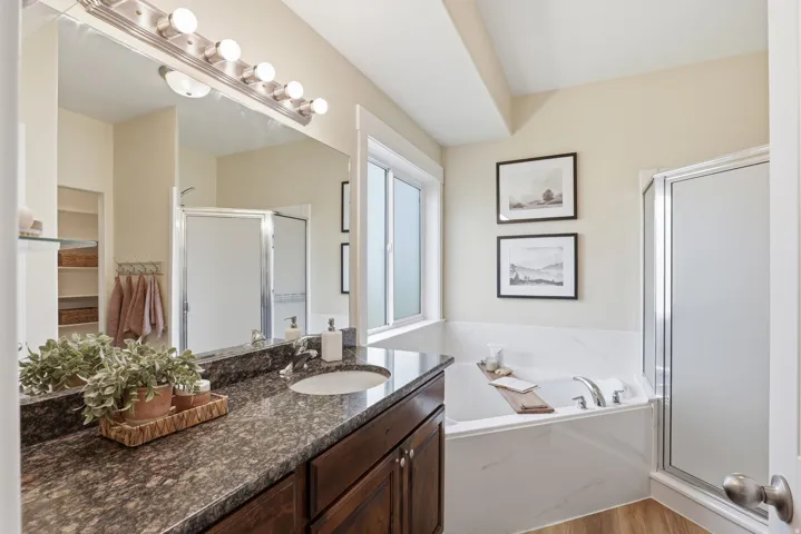 Bathroom featuring a double vanity with dark wood cabinetry, a large built-in mirror with strip lighting, a soaking tub with white tiled surround, a separate shower with a frosted glass door, and light wood-finish flooring