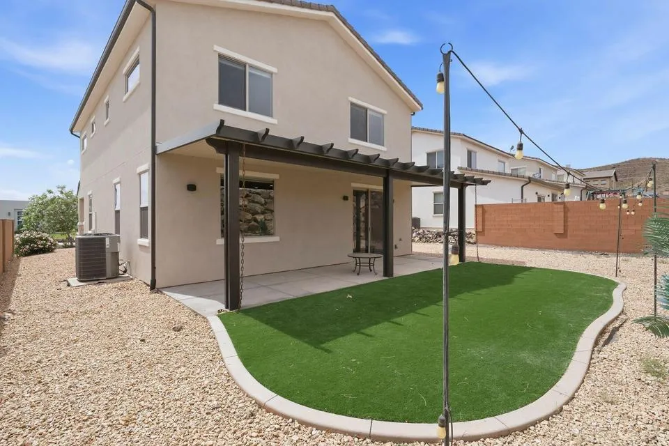Expansive backyard featuring a covered patio, artificial turf lawn, decorative gravel landscaping, string lighting, and a block wall perimeter