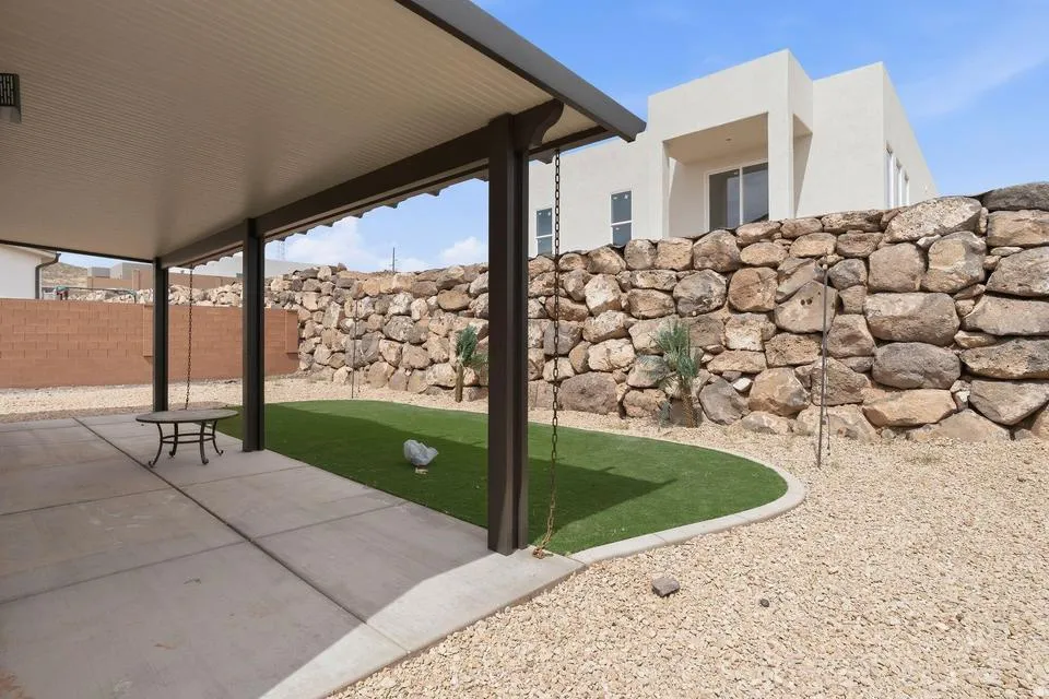 Covered patio with concrete flooring, framed by a stone retaining wall and artificial turf area