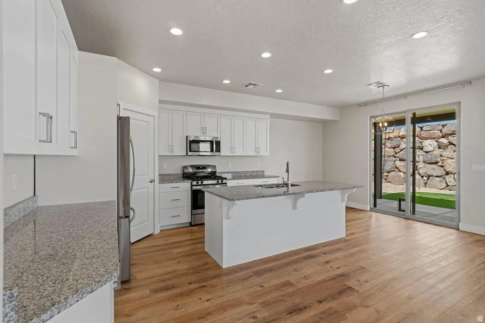 Modern kitchen featuring white cabinetry, granite-finish countertops, stainless steel appliances, a central island with sink, and wood-finish flooring