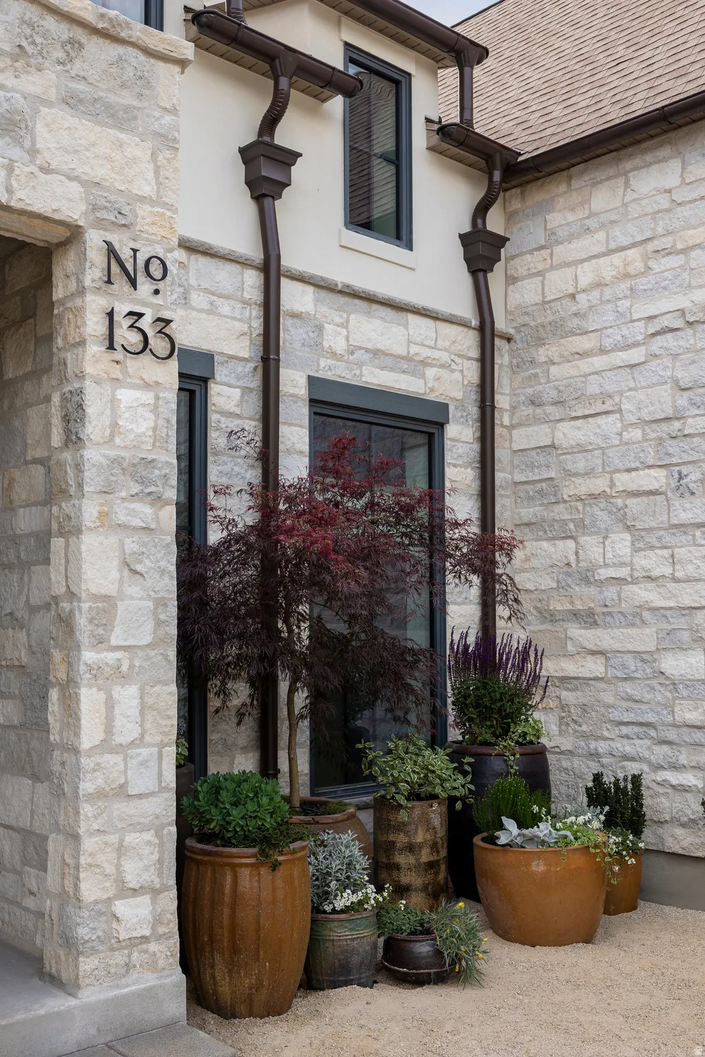 Stone facade featuring a prominent house number, dark-framed windows, decorative downspouts, and an assortment of potted plants on a gravel ground cover