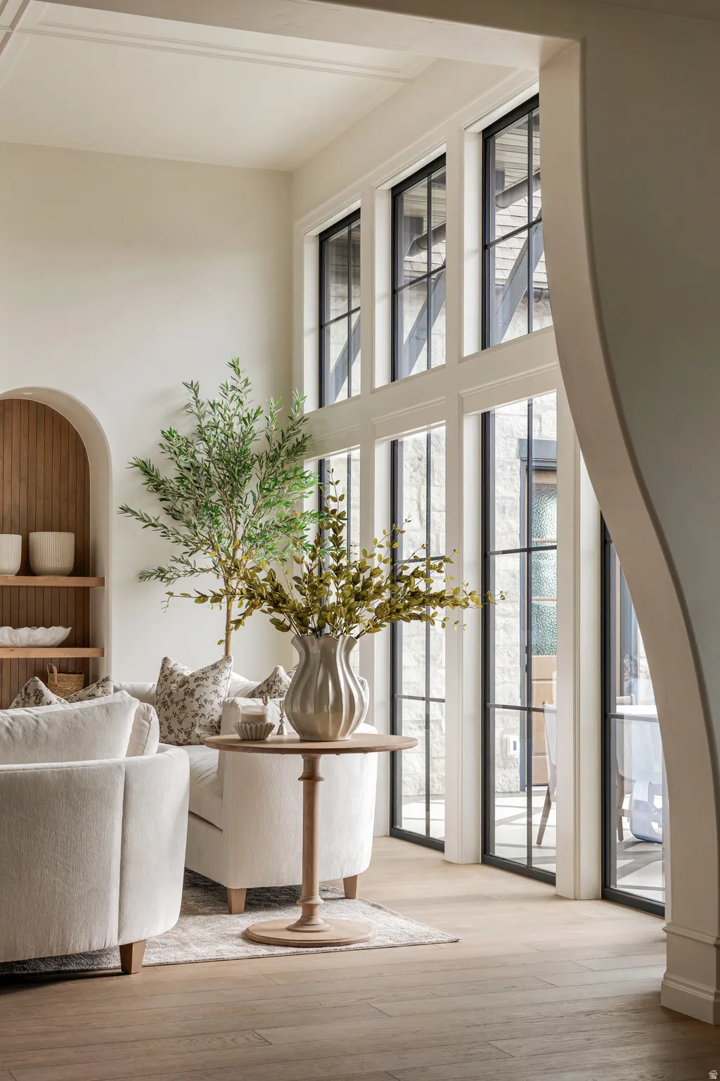 Expansive windows with black framing, wood-finish flooring, high ceilings, and an arched built-in shelving unit with wood paneling