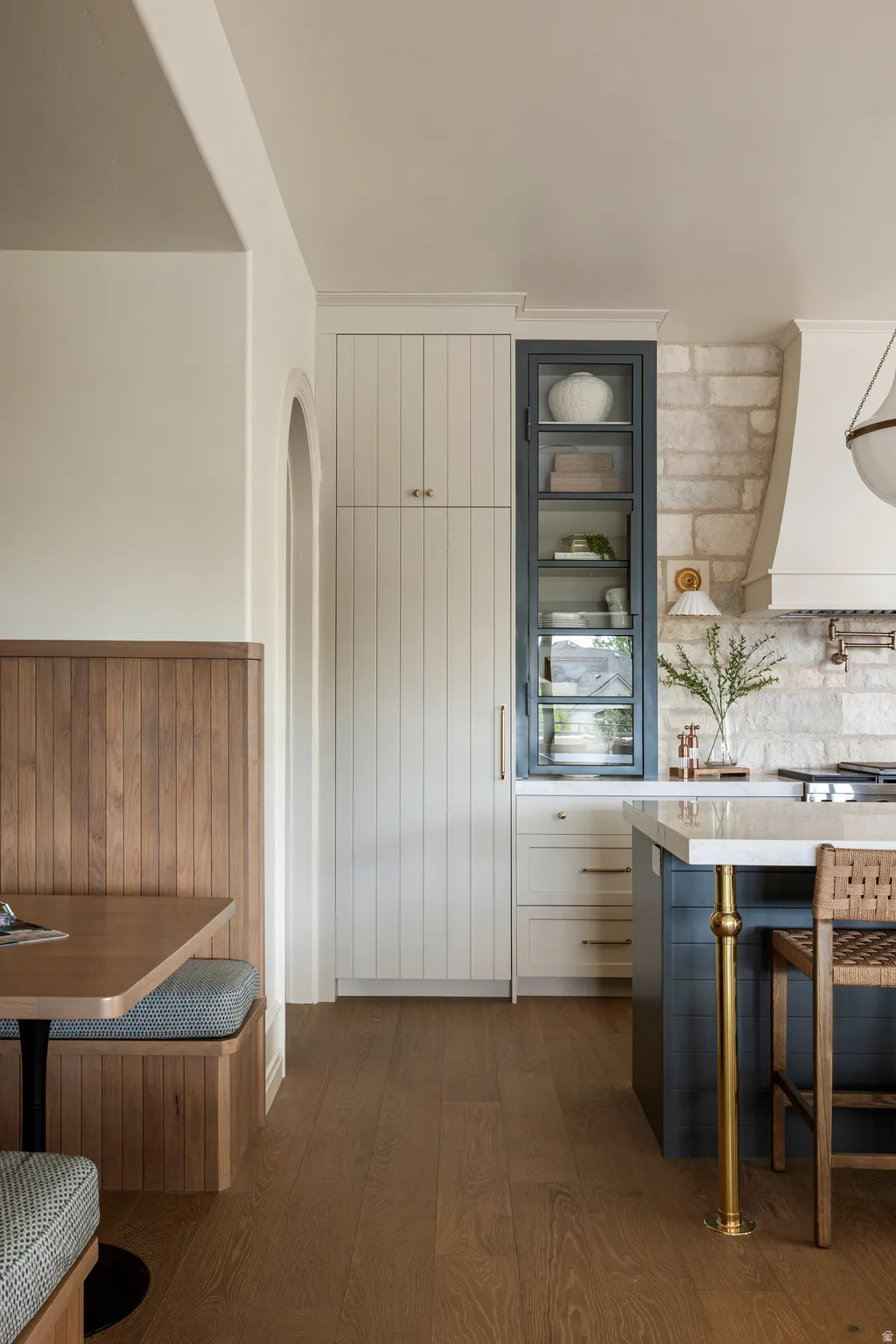 Open-concept kitchen featuring wood-finish flooring, a stone accent wall, white cabinetry with brass hardware, a dark-toned island with a light-toned countertop, and a built-in banquette with wood paneling