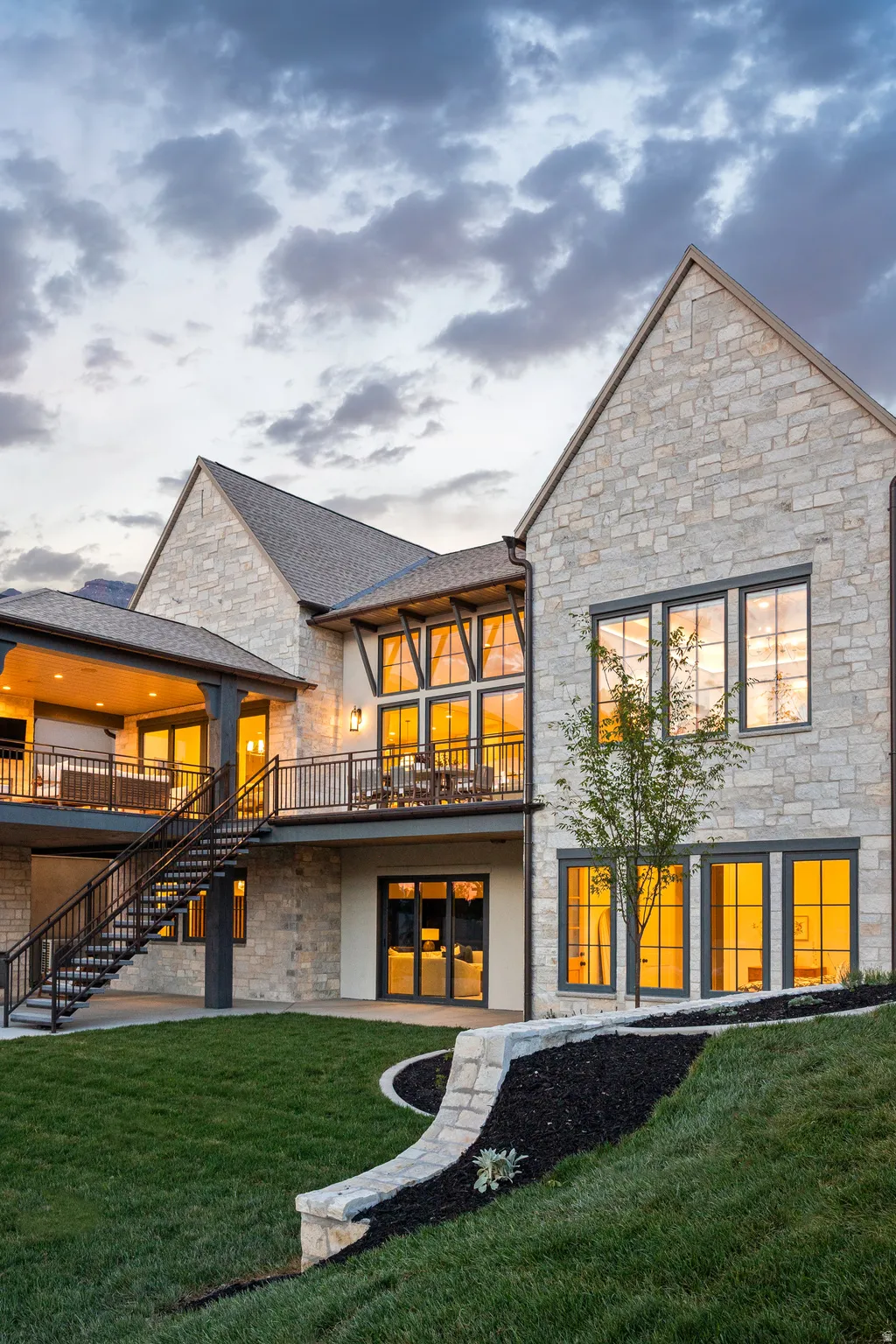 Stone facade residence featuring a covered balcony, an open deck, a metal staircase, and a terraced lawn with a stone retaining wall