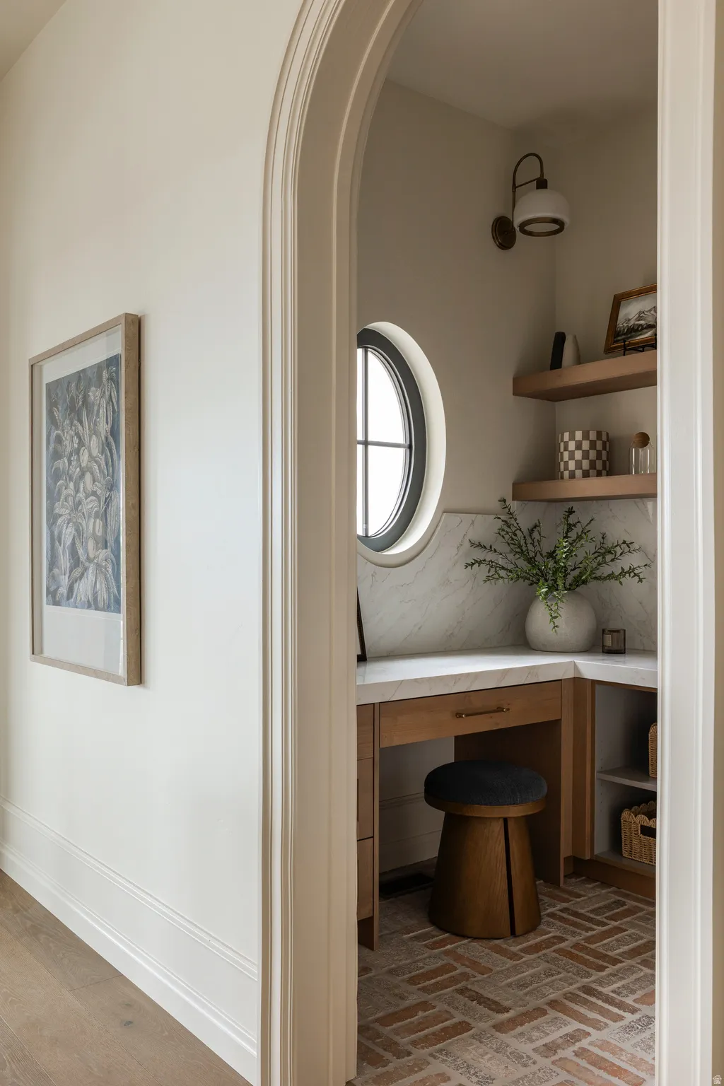 Arched doorway entrance leading to a dedicated workspace featuring a round window, built-in desk with a stone backsplash, integrated shelving, and herringbone brick flooring