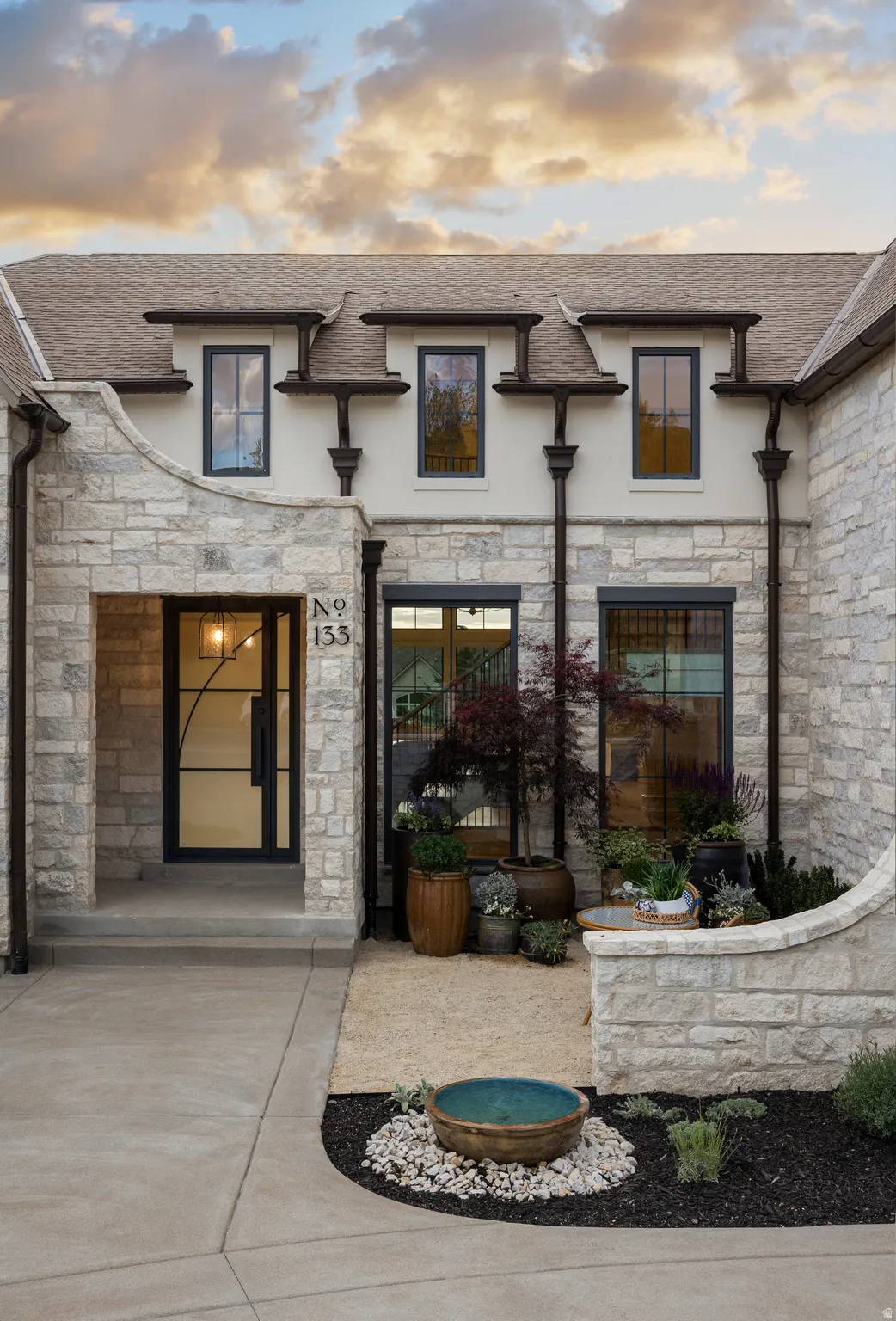 Stone facade exterior with dormer windows, a prominent entry door featuring decorative glass and metalwork, and a landscaped courtyard with a water feature
