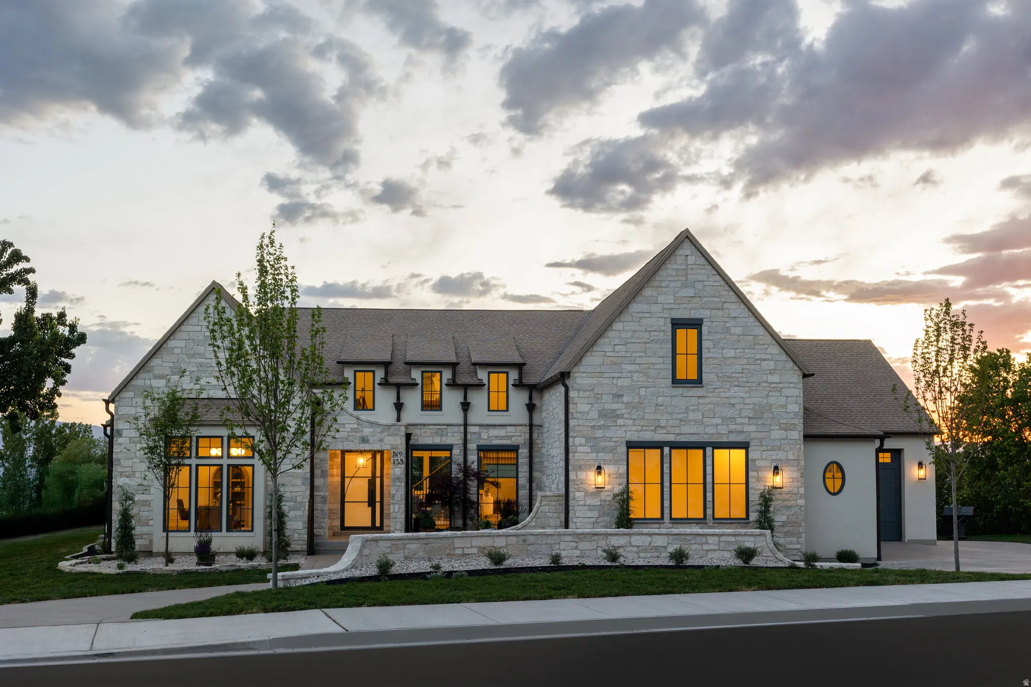 Stone façade residence featuring a multi-gabled roofline, prominent black-framed windows, a stucco section with an oval window, and a paved walkway