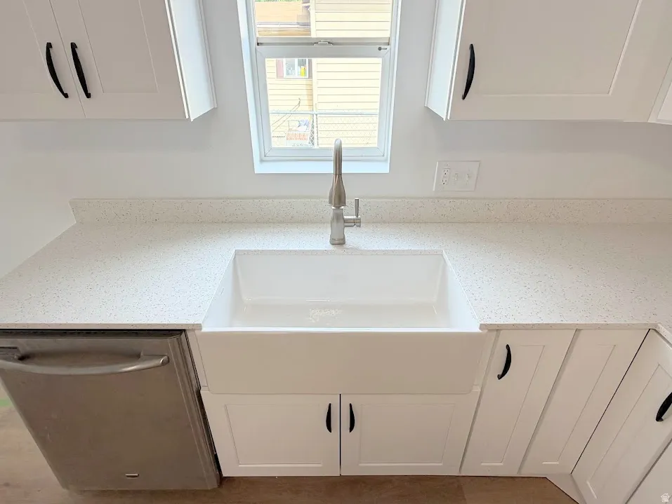 White kitchen featuring a farmhouse sink, stainless steel faucet, white shaker cabinetry with matte black hardware, and speckled light-toned countertops