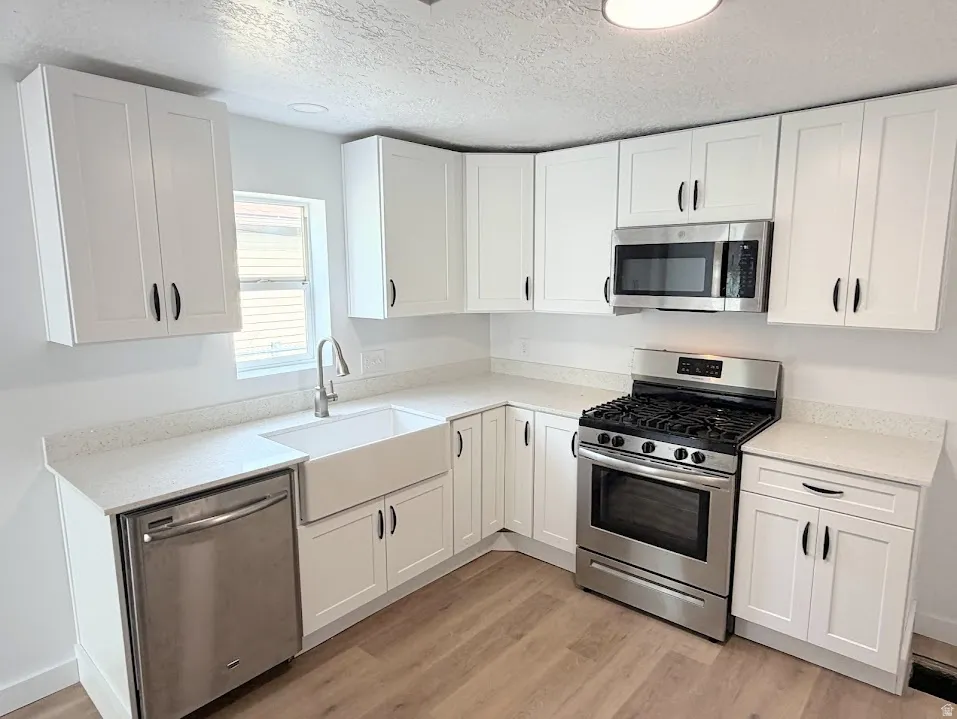 Kitchen featuring white shaker cabinetry, light-toned countertops, stainless steel appliances, a farmhouse-style sink, and wood-finish flooring