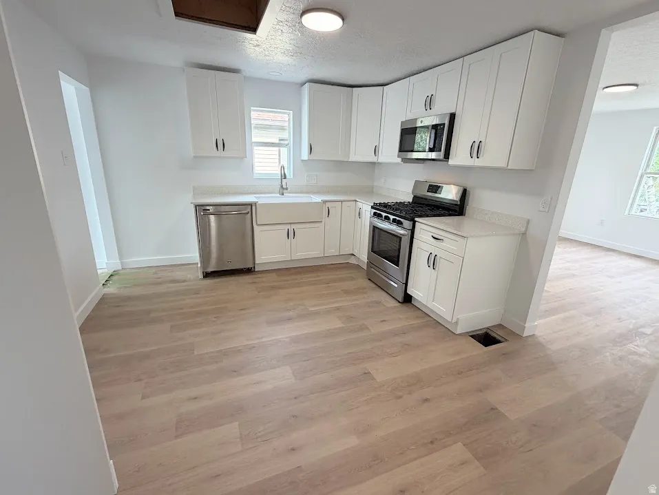 Bright kitchen featuring white shaker cabinetry, stainless steel appliances, a farmhouse sink, light-toned wood-finish flooring, and light-colored countertops