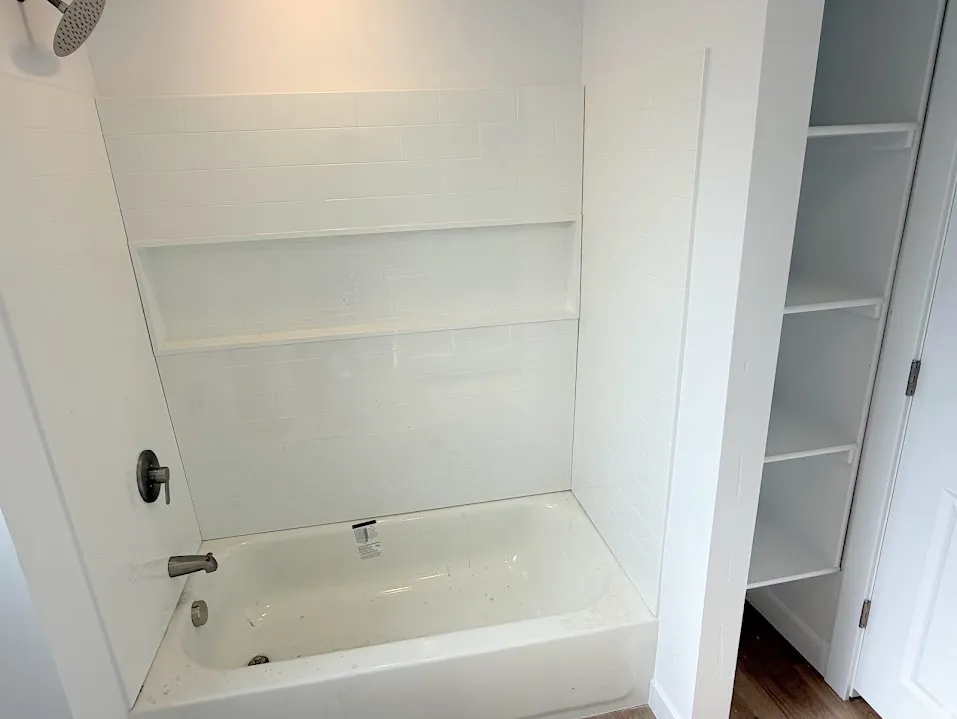 Bathtub and shower combination featuring white subway tile surround with two recessed niches, a dark bronze shower head and faucet, and a adjacent built-in shelving unit